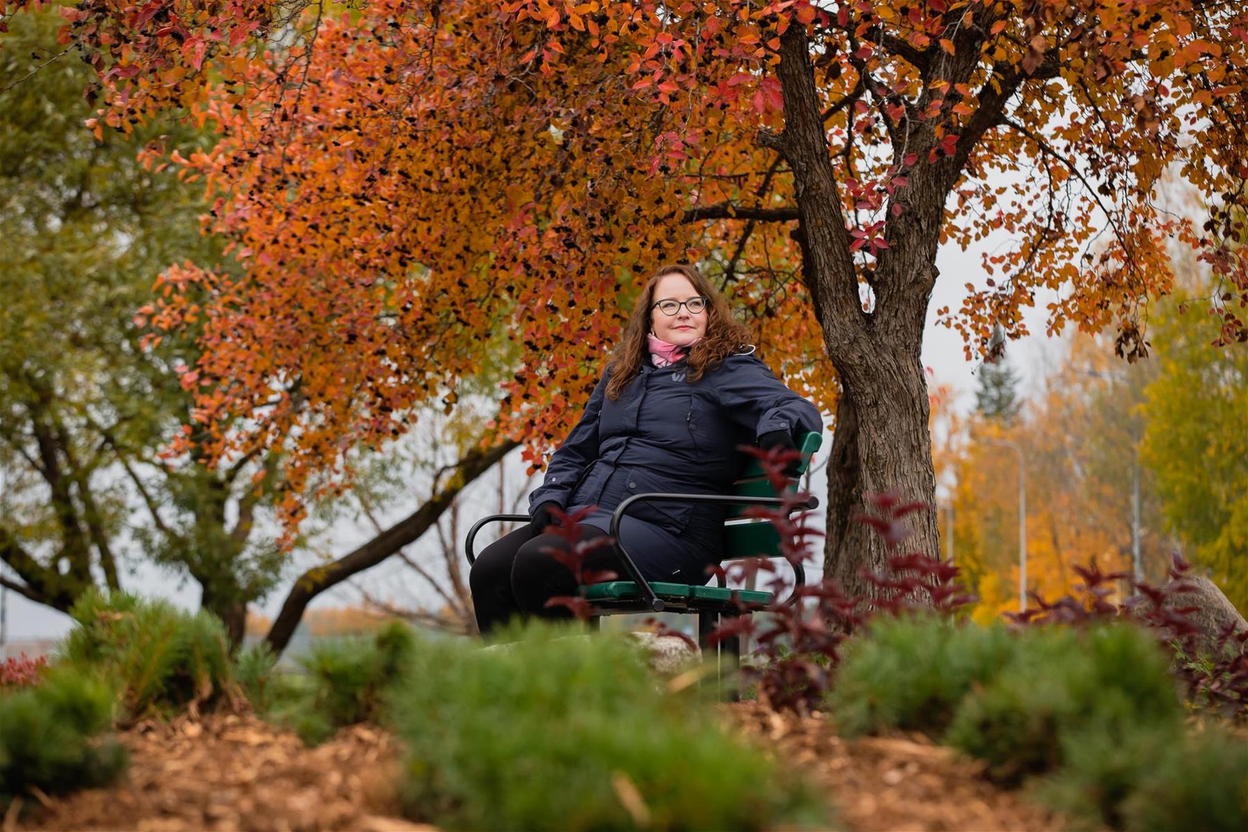 Kaupunginpuutarhuri Laura Ratilainen istuu penkillä puistossa, kuvassa puita ja viheristutusta